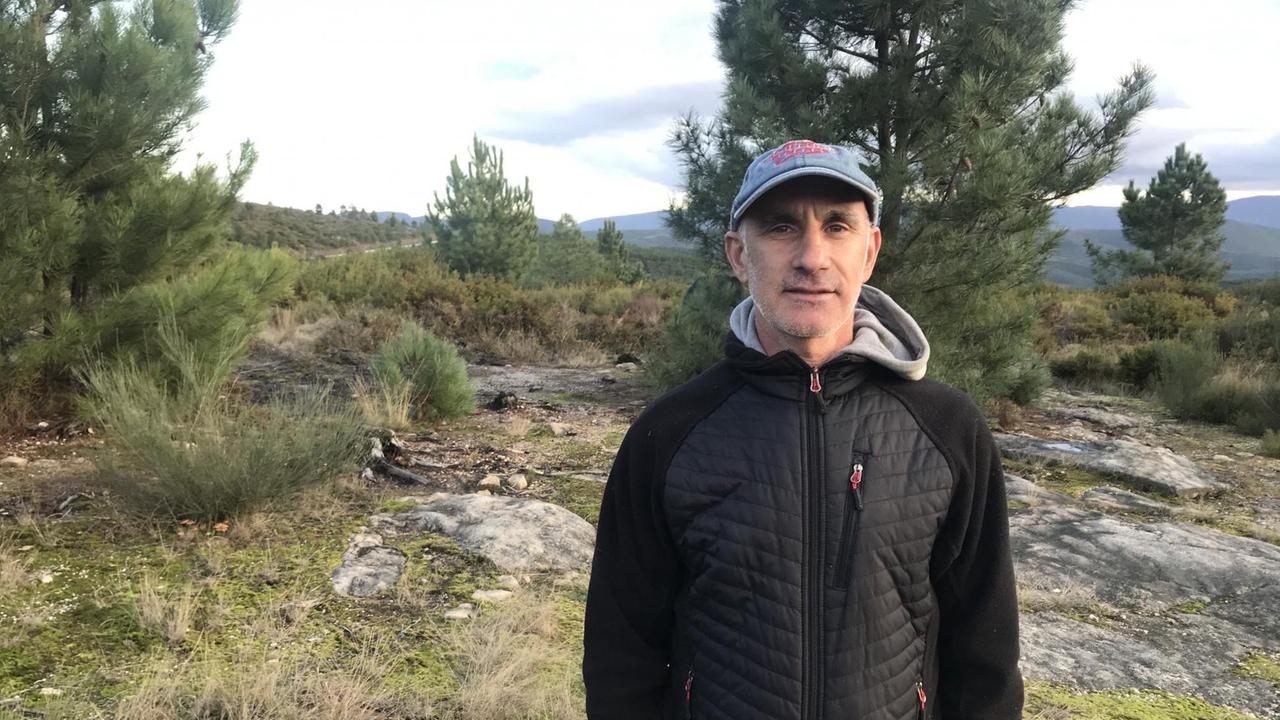 Ein älterer schmaler Mann mit Käppi und Anorak steht vor einer Landschaft mit Tannen, großen flachen Steinen und Hügeln im Hintergrund und schaut entschlossen in die Kamera. Ein älterer schmaler Mann mit Käppi und Anorak steht vor einer Landschaft mit Tannen, großen flachen Steinen und Hügeln im Hintergrund und schaut entschlossen in die Kamera.