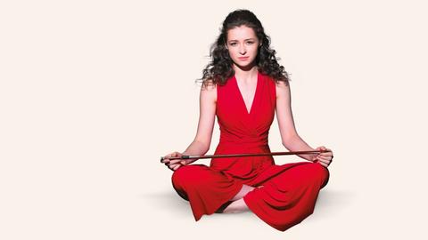 Die Musikerin sitzt ganz in Rot auf einem weißen Boden, ihren Bogen quer über die Knie gelegt.