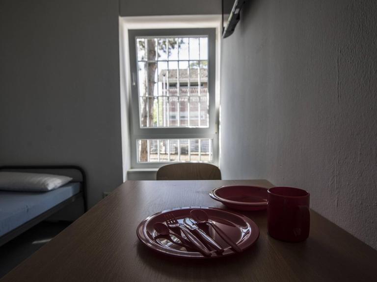 Ein Zimmer in einem Abschiebegefängnis mit einem Tisch und einem Bett vor einem vergitterten Fenster.