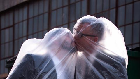 Die beiden Musiker, jeweils mit einer Plastiktüte über dem Kopf, küssen sich.