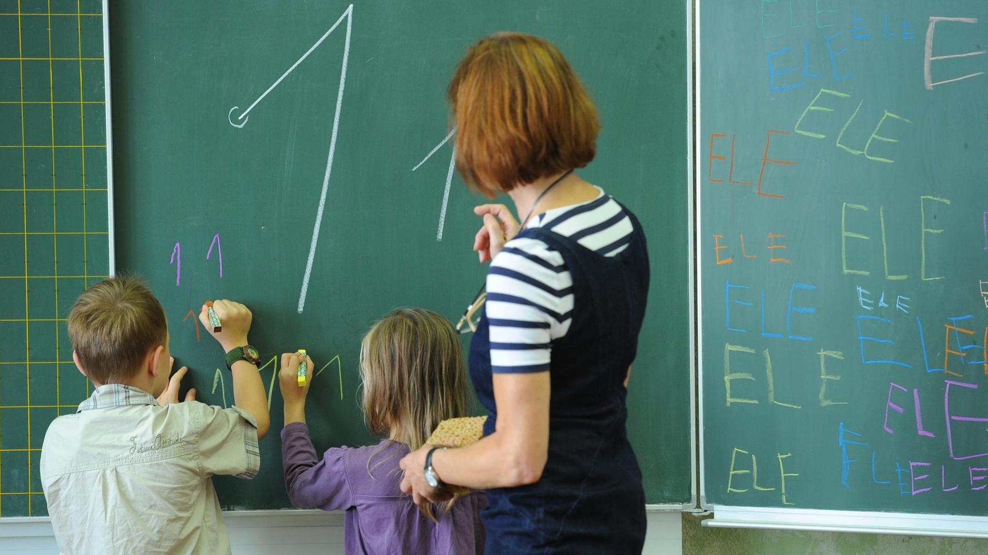 Zwei Kinder schreiben unter Anleitung ihrer Lehrerin an die Tafel.