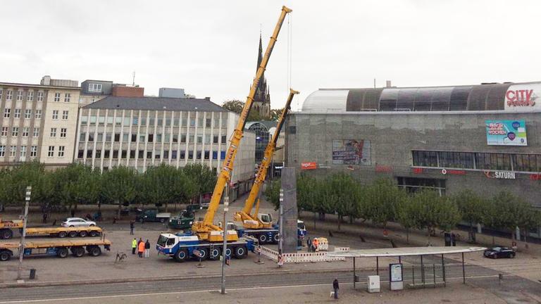 Ein großer Stadtplatz auf dem Kräne einen Obelisken abbauen Ein großer Stadtplatz auf dem Kräne einen Obelisken abbauen