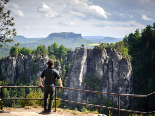 Ein Wanderer steht am 19.05.2015 in der Nähe von Rathen (Sachsen) auf einem Felsen in der Nationalparkregion Sächsische Schweiz und schaut zur Bastei hinüber, im Hintergrund erhebt sich der Lilienstein.