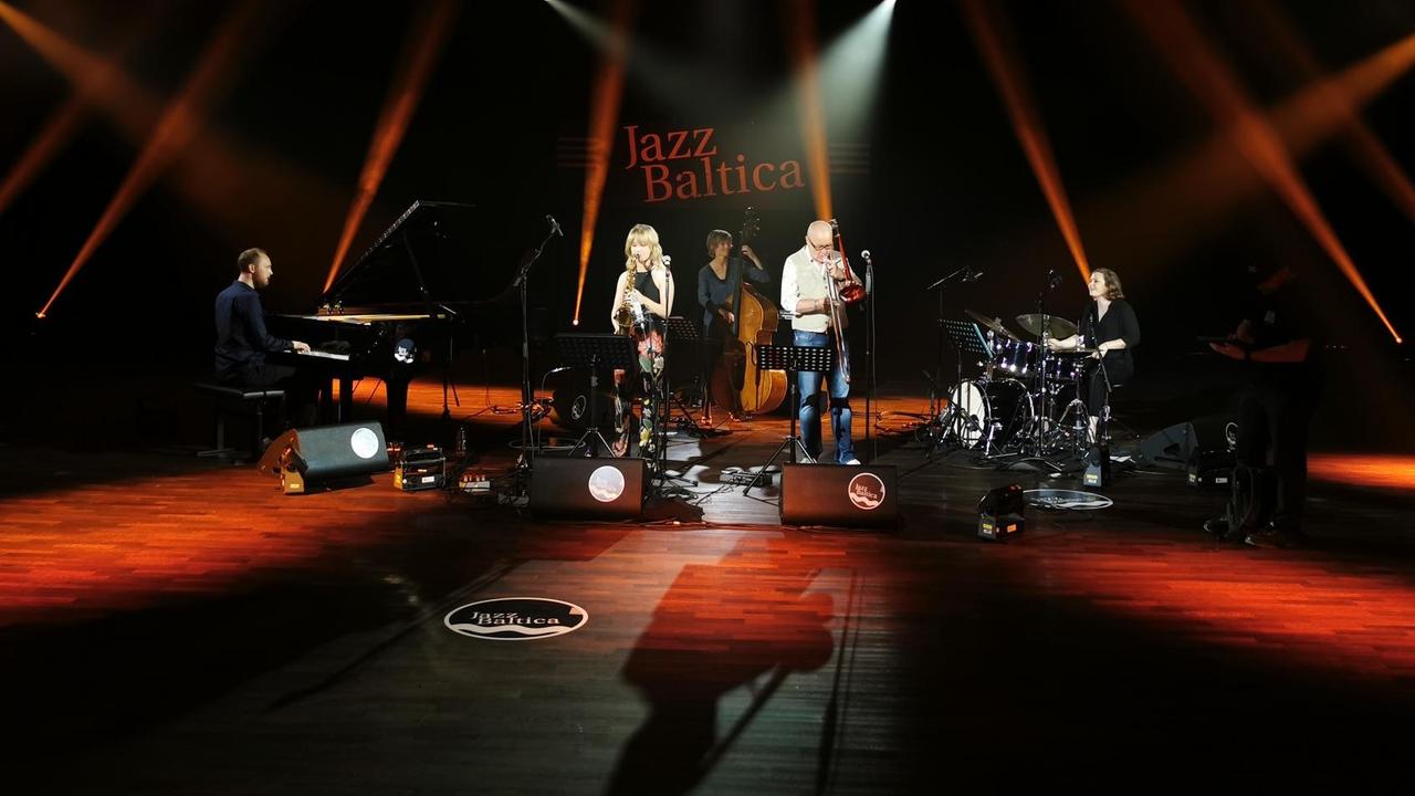 Fünf Musikerinnen und Musiker spielen auf einer in rot ausgeleuchteten Bühne beim Festival Jazzbaltica Fünf Musikerinnen und Musiker spielen auf einer in rot ausgeleuchteten Bühne beim Festival Jazzbaltica