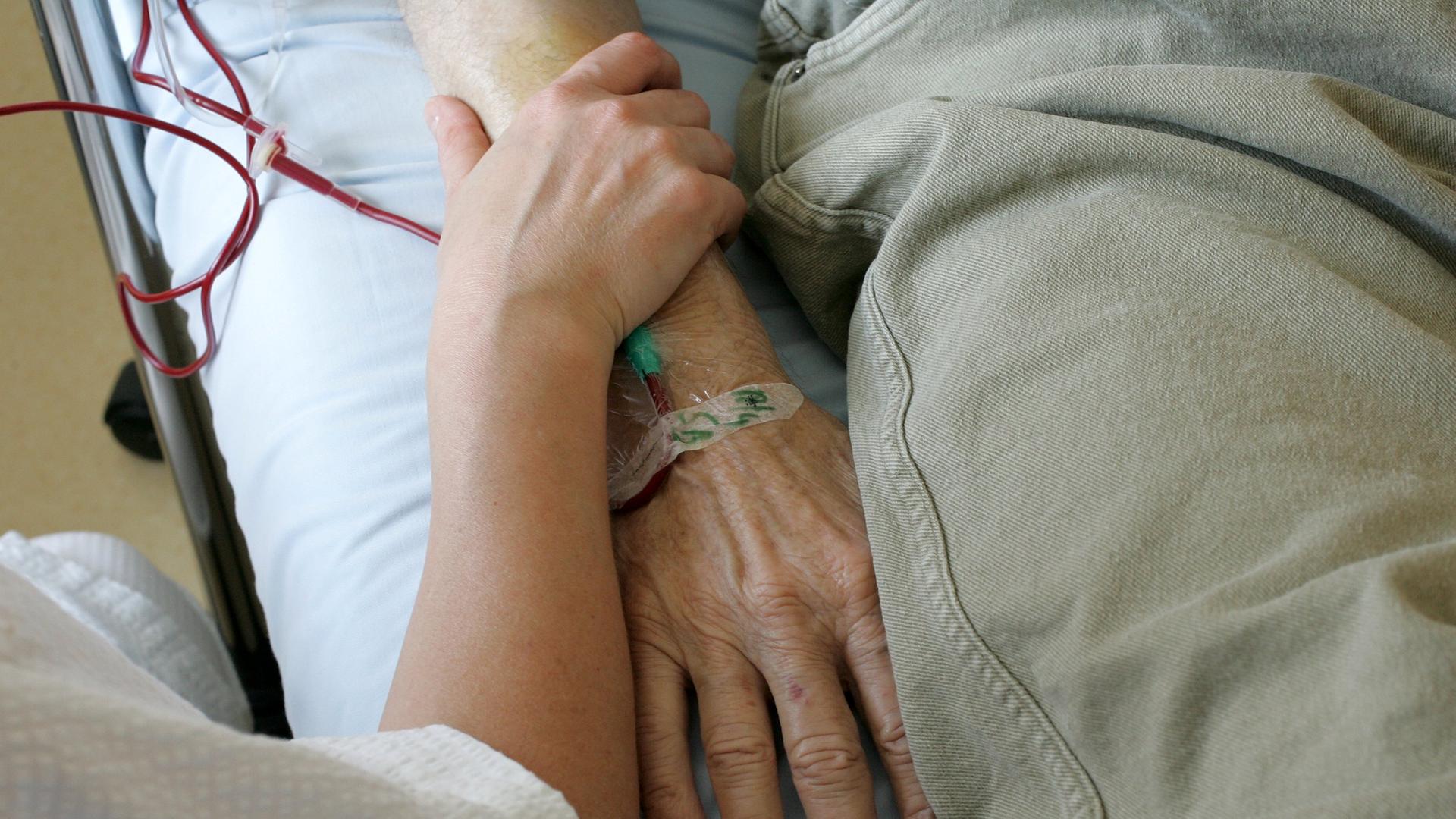 Ein jüngerer Mensch umfasst das Armgelenk einer älteren Person, die im Krankenbett liegt. Ein jüngerer Mensch umfasst das Armgelenk einer älteren Person, die im Krankenbett liegt.