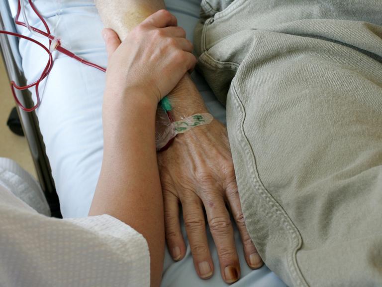 Ein jüngerer Mensch umfasst das Armgelenk einer älteren Person, die im Krankenbett liegt.