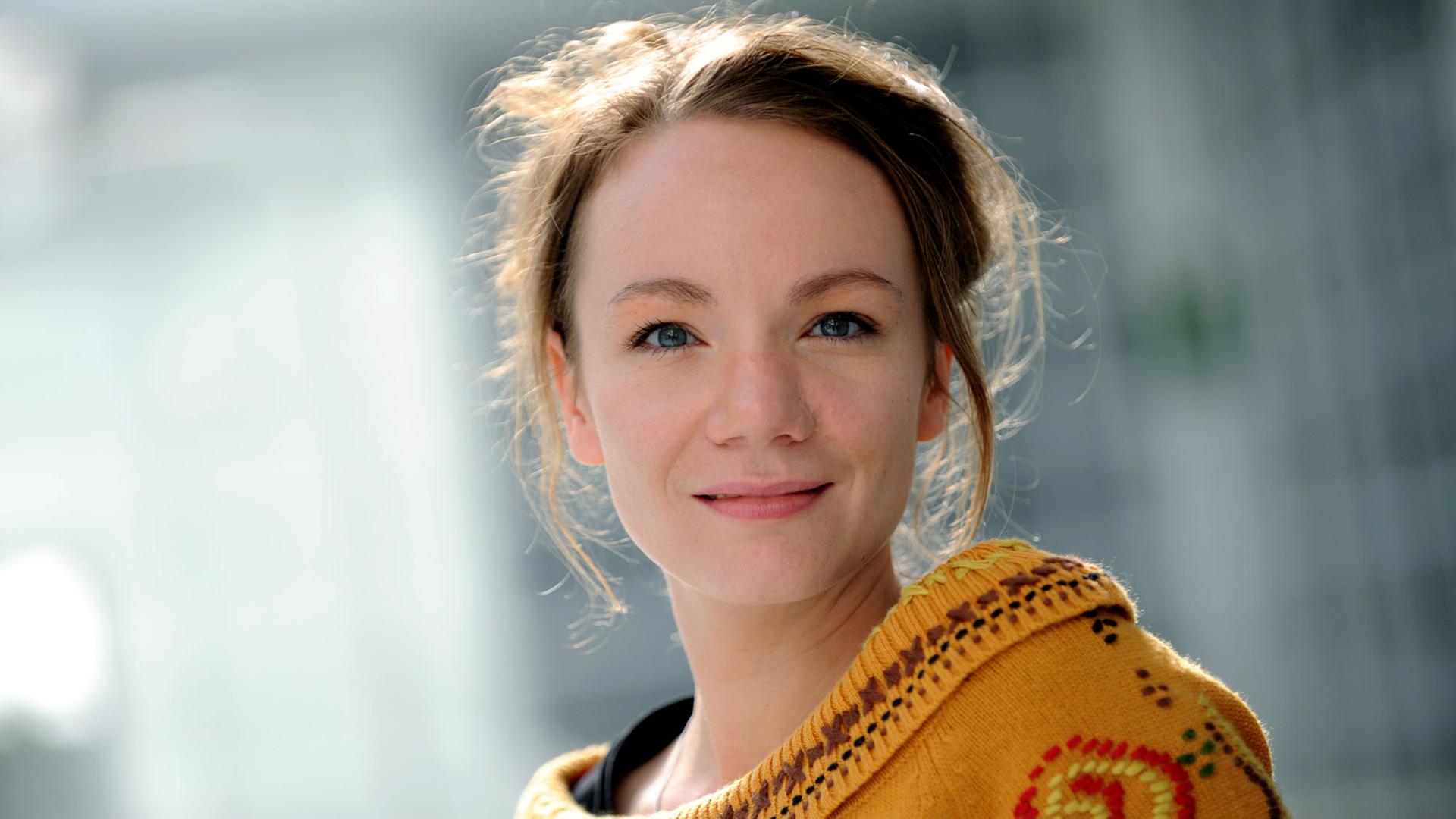 Porträtaufnahme einer jungen Frau, die einen gelben Pullover trägt und in die Kamera lächelt.