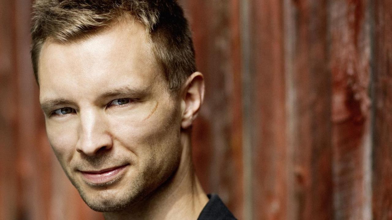 Ein Mann mit markanten Gesichtszügen steht vor einer Holzwand Ein Mann mit markanten Gesichtszügen steht vor einer Holzwand