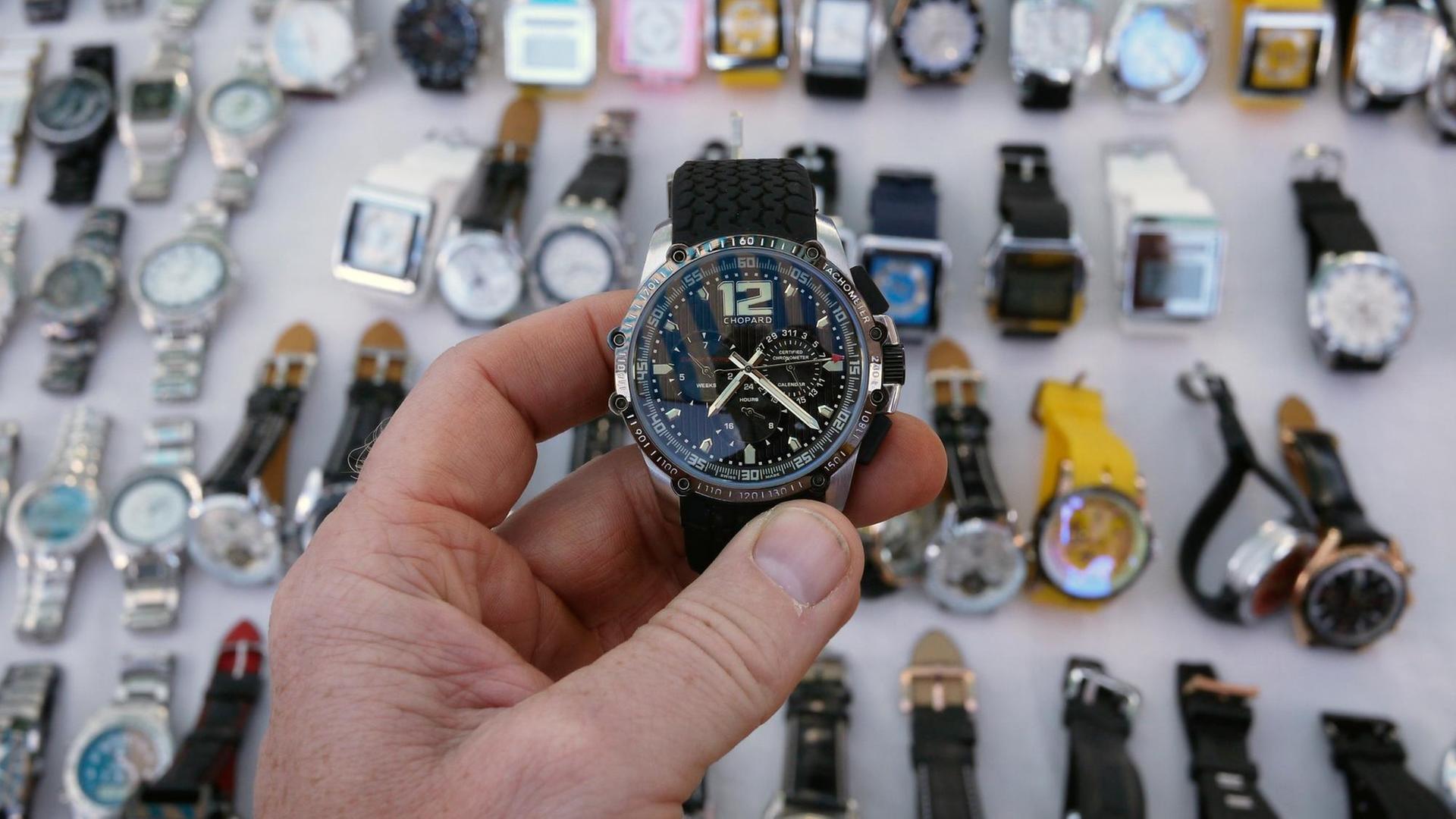 Eine Hand hält eine plagiierte Uhr der Marke Chopard in die Kamera, darunter sind in einer Auslage zahlreiche weiterer nachgemachte Uhren zu sehen Eine Hand hält eine plagiierte Uhr der Marke Chopard in die Kamera, darunter sind in einer Auslage zahlreiche weiterer nachgemachte Uhren zu sehen