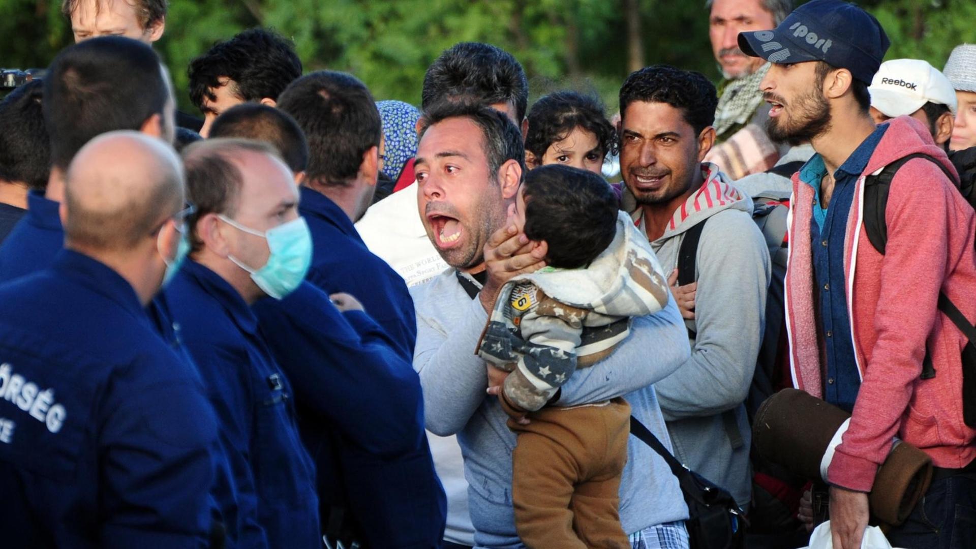 Polizisten in Uniform und zum Teil mit Mundschutz stehen aufgebrachten Flüchtlingen gegenüber. Ein Mann schreit die Polizisten an, hält ein Kind auf dem Arm. Polizisten in Uniform und zum Teil mit Mundschutz stehen aufgebrachten Flüchtlingen gegenüber. Ein Mann schreit die Polizisten an, hält ein Kind auf dem Arm.