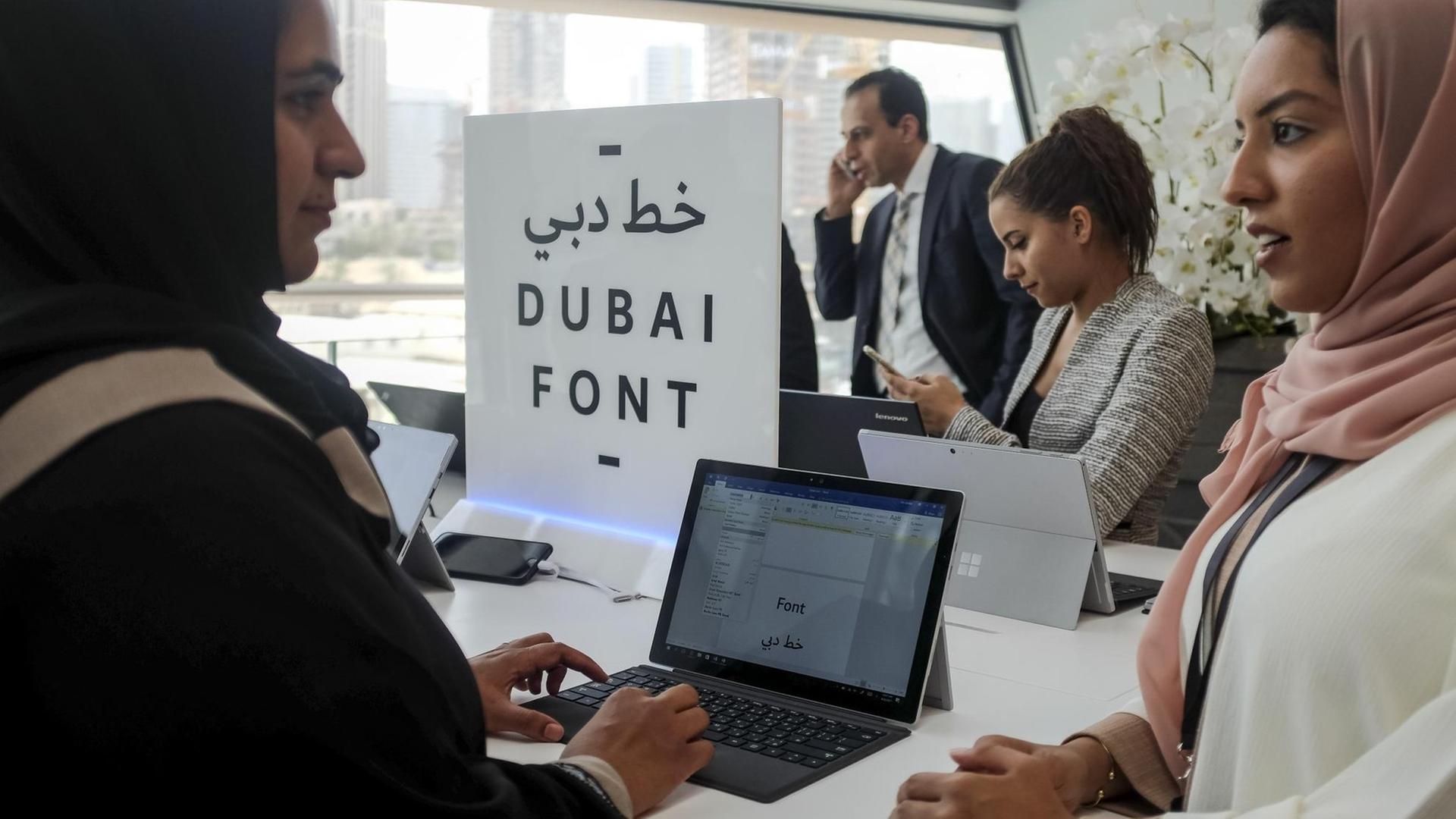 Sie sehen drei Frauenan Laptops, in der Mitte ein Schild mit der Aufschrift "DUBAI FONT". Im Hintergrund telefoniert ein Mann. Sie sehen drei Frauenan Laptops, in der Mitte ein Schild mit der Aufschrift "DUBAI FONT". Im Hintergrund telefoniert ein Mann.