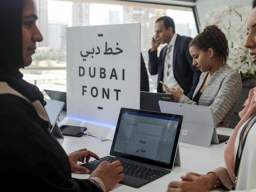 Sie sehen drei Frauenan Laptops, in der Mitte ein Schild mit der Aufschrift "DUBAI FONT". Im Hintergrund telefoniert ein Mann.