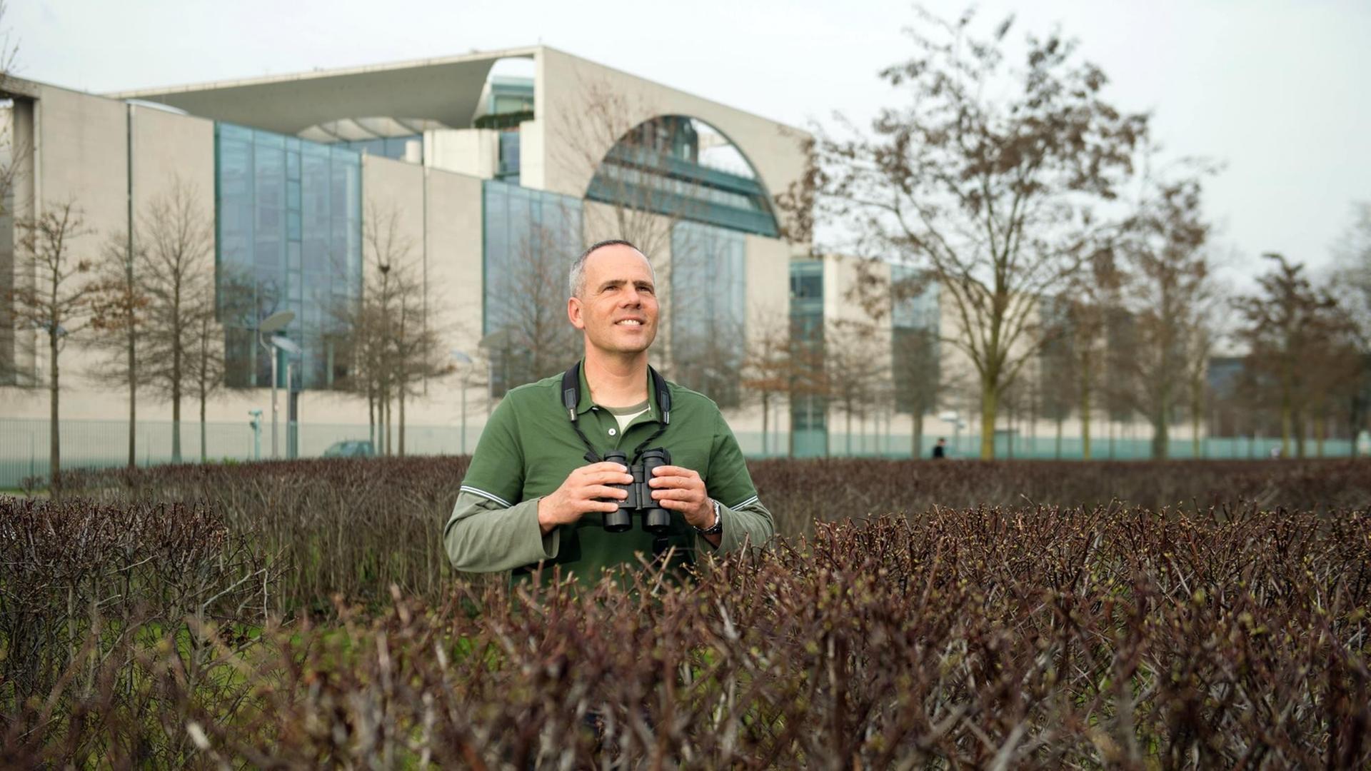 Der Berliner Wildtierbeauftragte Derk Ehlert steht mit einem Fernglas um den Hals zwischen Sträuchern im Berliner Tiergarten, im Hintergrund das Bundeskanzleramt. Der Berliner Wildtierbeauftragte Derk Ehlert steht mit einem Fernglas um den Hals zwischen Sträuchern im Berliner Tiergarten, im Hintergrund das Bundeskanzleramt.