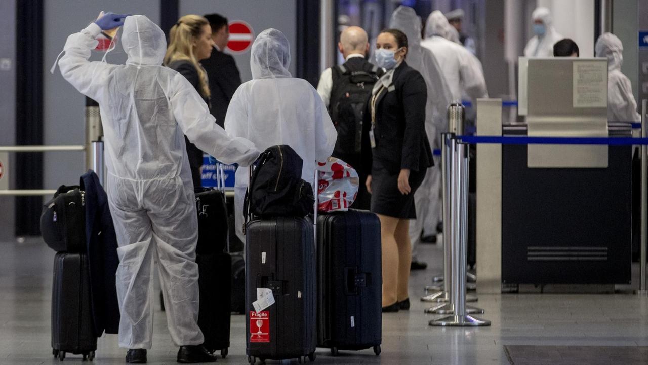 Die Reisenden mit Koffern sind von hinten und aus größerer Entfernung fotografiert. Es stehen auch Reisende ohne Schutzanzüge sowie Sicherheitspersonal mit Schutzmasken dort. Die Reisenden mit Koffern sind von hinten und aus größerer Entfernung fotografiert. Es stehen auch Reisende ohne Schutzanzüge sowie Sicherheitspersonal mit Schutzmasken dort.