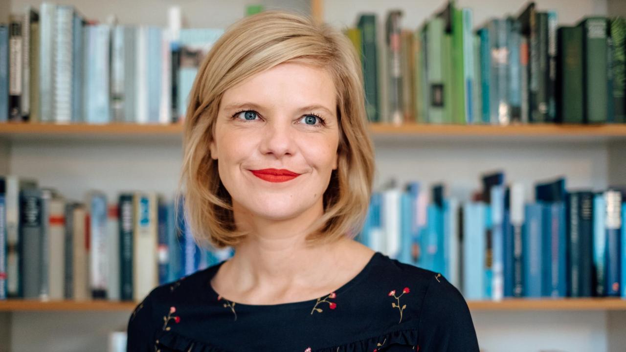 Eine blonde Frau mit rotem Lippenstift steht vor einem Bücherregal. Eine blonde Frau mit rotem Lippenstift steht vor einem Bücherregal.