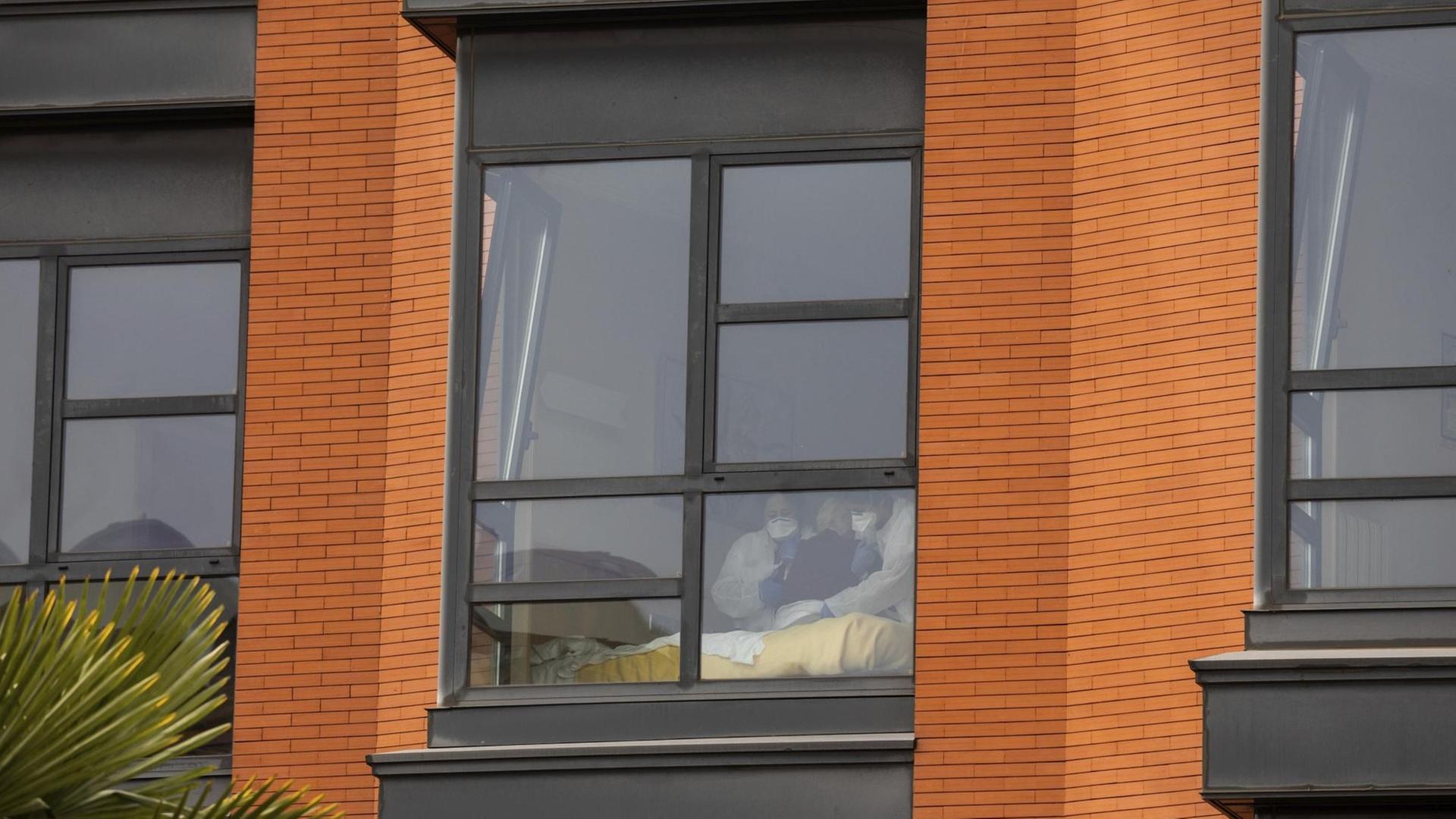 Man sieht die beiden Pflegekräfte und die Seniorin undeutlich durch ein Fenster an der Fassade. Daneben zwei weitere Fenster. Man sieht die beiden Pflegekräfte und die Seniorin undeutlich durch ein Fenster an der Fassade. Daneben zwei weitere Fenster.