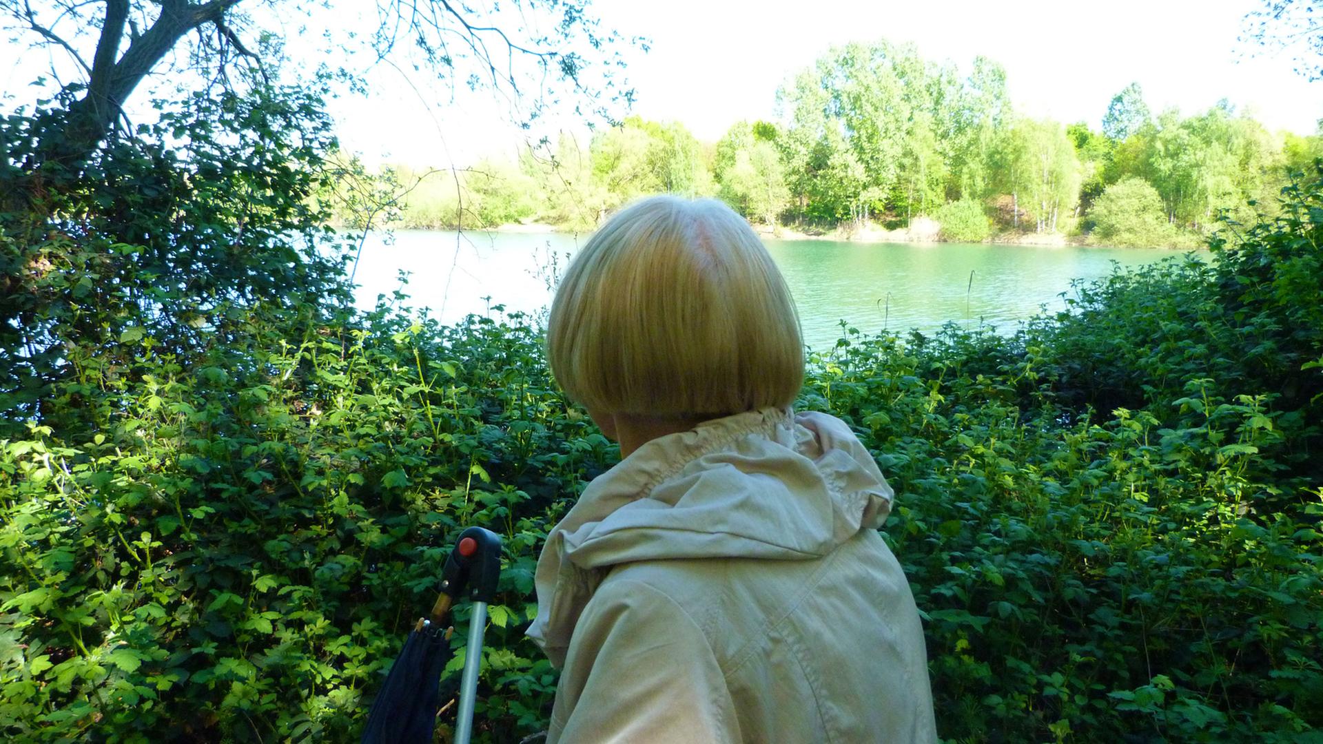 Margot an einem bedeutenden Ort ihres Lebens. Zu sehen: Margot mit dem Rücken zur Kamera. Sie hat halblange blonde Haare, trägt einen grauen Anorak und schaut auf einen See. Margot an einem bedeutenden Ort ihres Lebens. Zu sehen: Margot mit dem Rücken zur Kamera. Sie hat halblange blonde Haare, trägt einen grauen Anorak und schaut auf einen See.