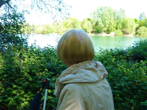 Margot an einem bedeutenden Ort ihres Lebens. Zu sehen: Margot mit dem Rücken zur Kamera. Sie hat halblange blonde Haare, trägt einen grauen Anorak und schaut auf einen See.