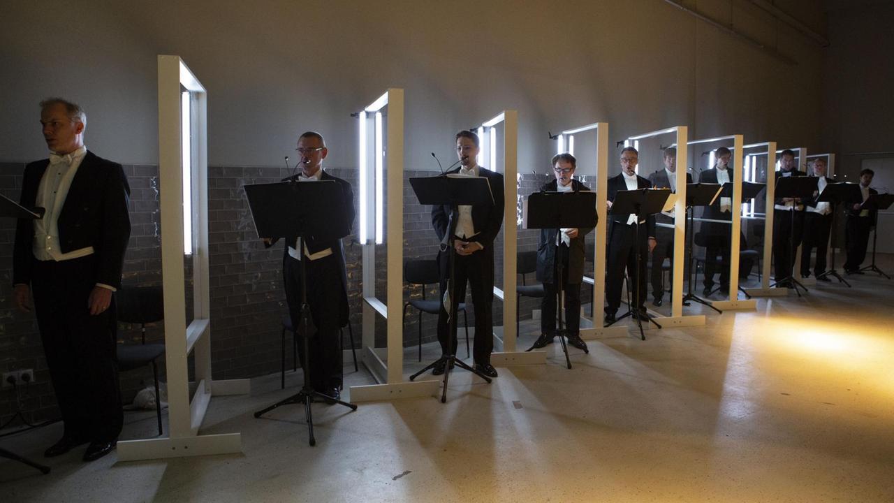Sänger des Chores stehen zwischen Plexiglaswänden in einer geheimnisvoll ausgeleuchteten Industriehalle.
