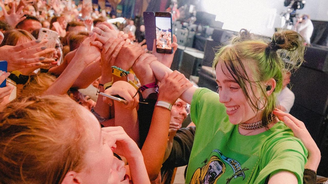 Billie Eilish steht mit ausgebreiteten Armen ganz nah an ihren Fans, die ihre Hände und Arme berühren. Billie Eilish steht mit ausgebreiteten Armen ganz nah an ihren Fans, die ihre Hände und Arme berühren.