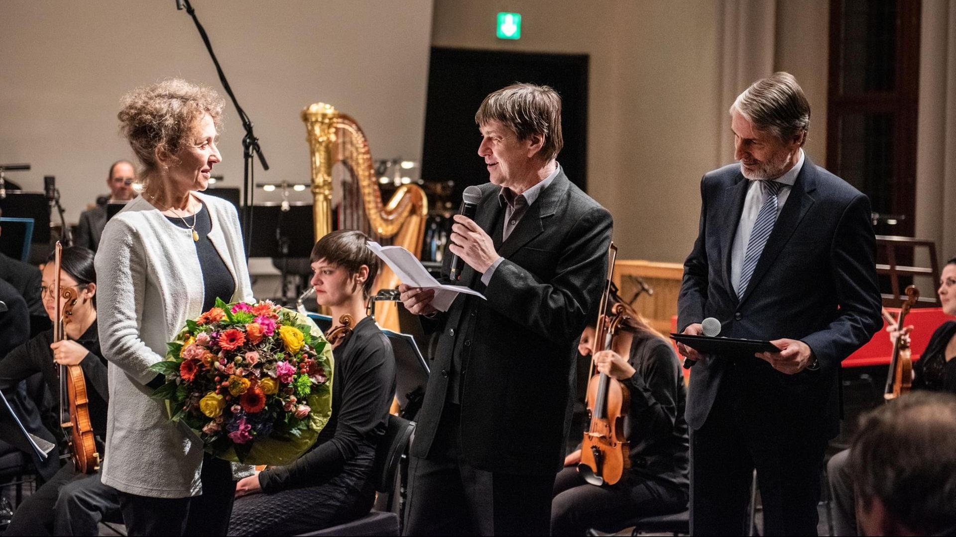 Links hält eine Frau einen Blumenstrauß in der Hand, rechts von ihr stehen zwei Herren im Anzug mit Mikrofon in der Hand, einer davon hält eine Ansprache. Im Hintergrund ist ein Orchester im Ausschnitt zu sehen. Links hält eine Frau einen Blumenstrauß in der Hand, rechts von ihr stehen zwei Herren im Anzug mit Mikrofon in der Hand, einer davon hält eine Ansprache. Im Hintergrund ist ein Orchester im Ausschnitt zu sehen.
