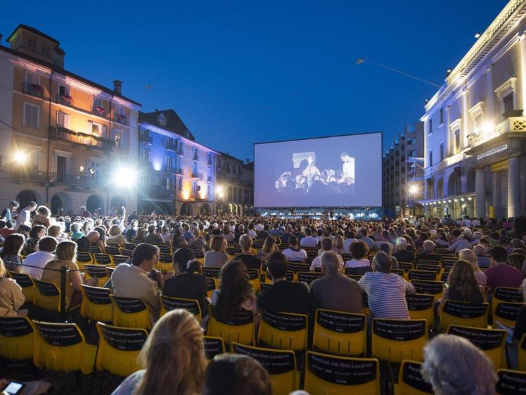 Zuschauer sitzen in der Nacht vor der offiziellen Eröffnung des Filmfestivals vor einer großen Leinwand mitten in Locarno
