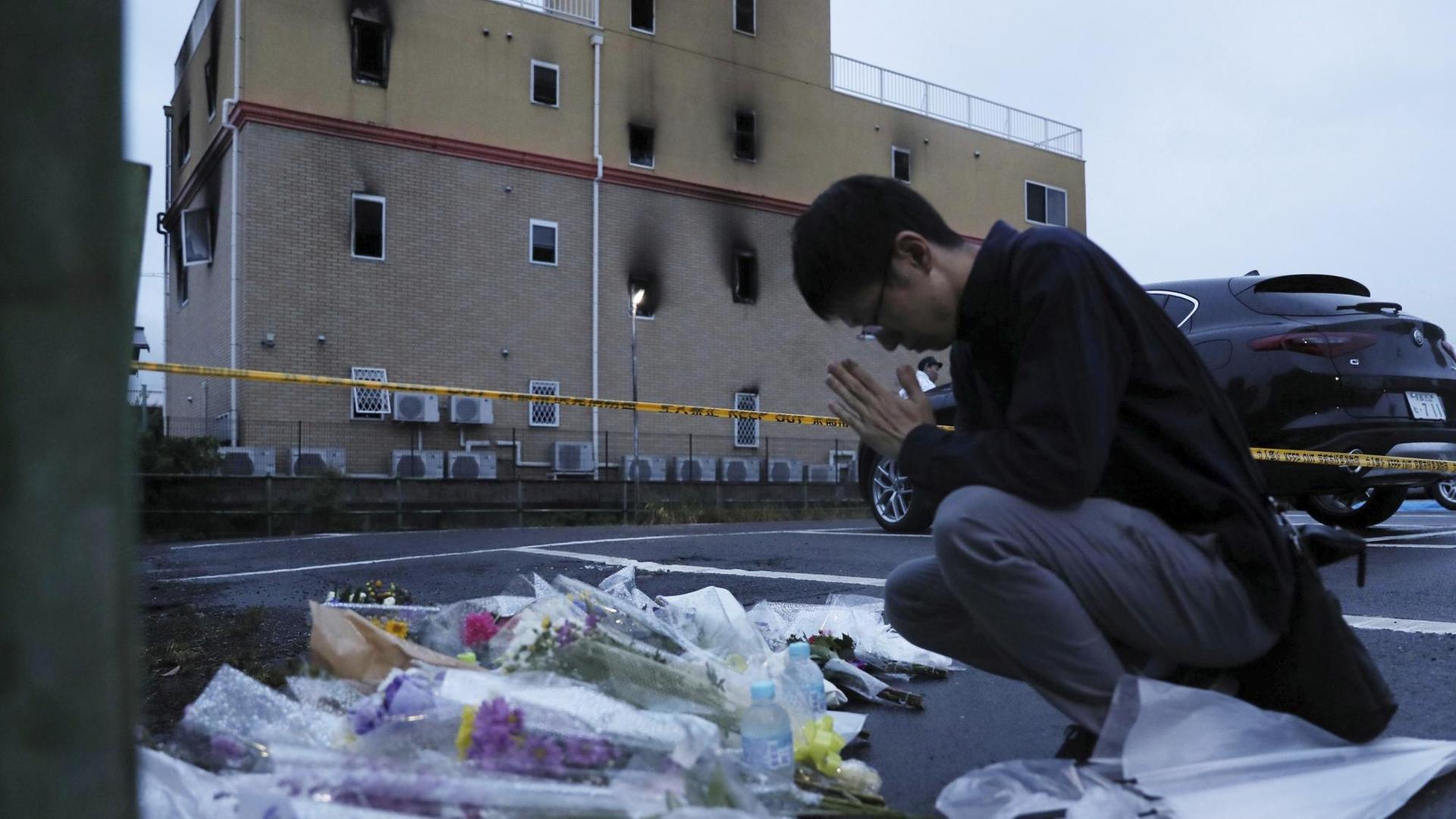 Ein Mann mit gefalteten Händen kniet vor dem Kyoto-Animation-Studio. Vor ihm liegen Blumen. Das Gebäude im Hintergrund hat Rußspuren. Ein Mann mit gefalteten Händen kniet vor dem Kyoto-Animation-Studio. Vor ihm liegen Blumen. Das Gebäude im Hintergrund hat Rußspuren.