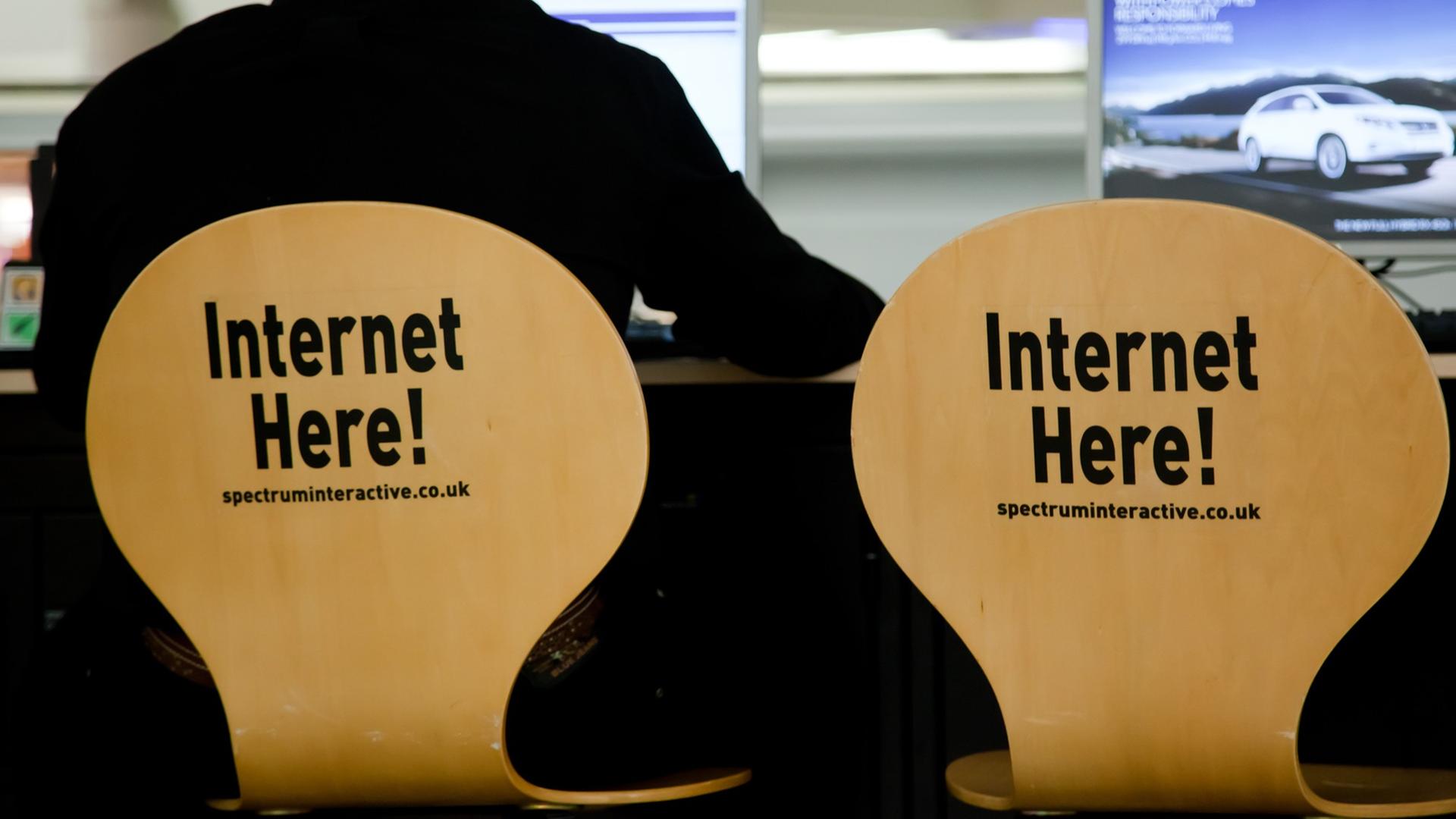 Ein Mann sitzt auf einem Stuhl, auf dessen Rückenlehne "Internet here!" steht, neben ihm ein identischer Stuhl und ein Computerbildschirm Ein Mann sitzt auf einem Stuhl, auf dessen Rückenlehne "Internet here!" steht, neben ihm ein identischer Stuhl und ein Computerbildschirm