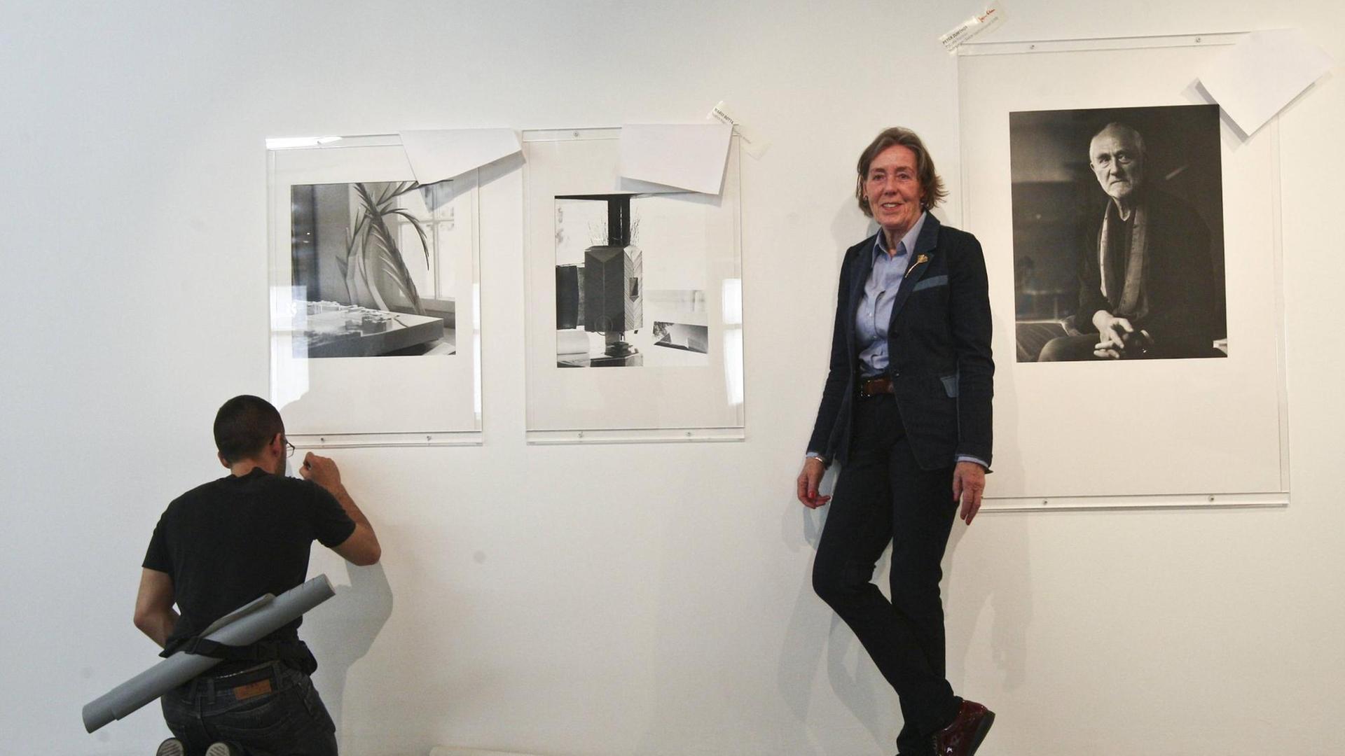 Ingrid von Kruse steht an eine Wand gelehnt. Ein Mann hockt auf dem Boden und besfstigt drei ihrer Fotografien an der Wand. Aufgenommen im April 2015 während der Vorbereitungen einer Ausstellung in Lissabon.