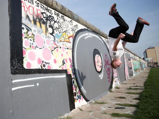 Ein junger Mann stößt sich artistisch von der Berliner East-Side-Gallery ab und macht einen Salto.