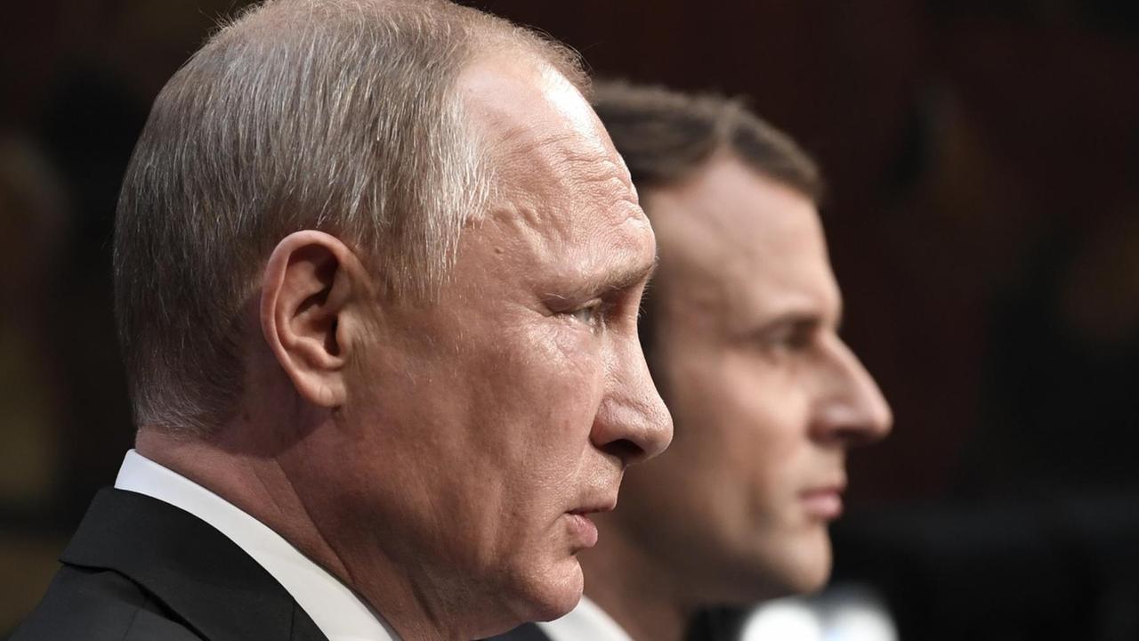 Nahaufnahme von Putins Kopf von der Seite, dahinter unscharf der Kopf Macrons vor einem dunklen Hintergrund. Nahaufnahme von Putins Kopf von der Seite, dahinter unscharf der Kopf Macrons vor einem dunklen Hintergrund.