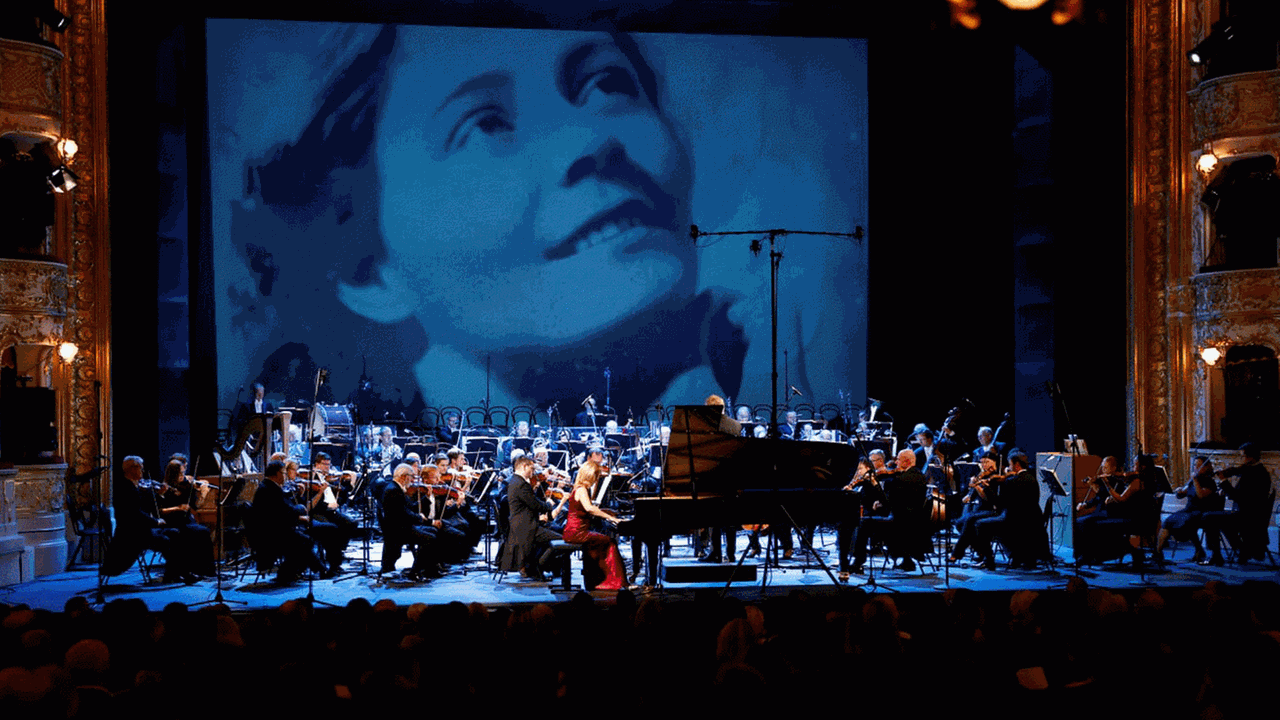 Das Bildnis von Vítězslava Kaprálová ist auf eine große Leinwand hinter dem Orchester sichtbar. Das Bildnis von Vítězslava Kaprálová ist auf eine große Leinwand hinter dem Orchester sichtbar.