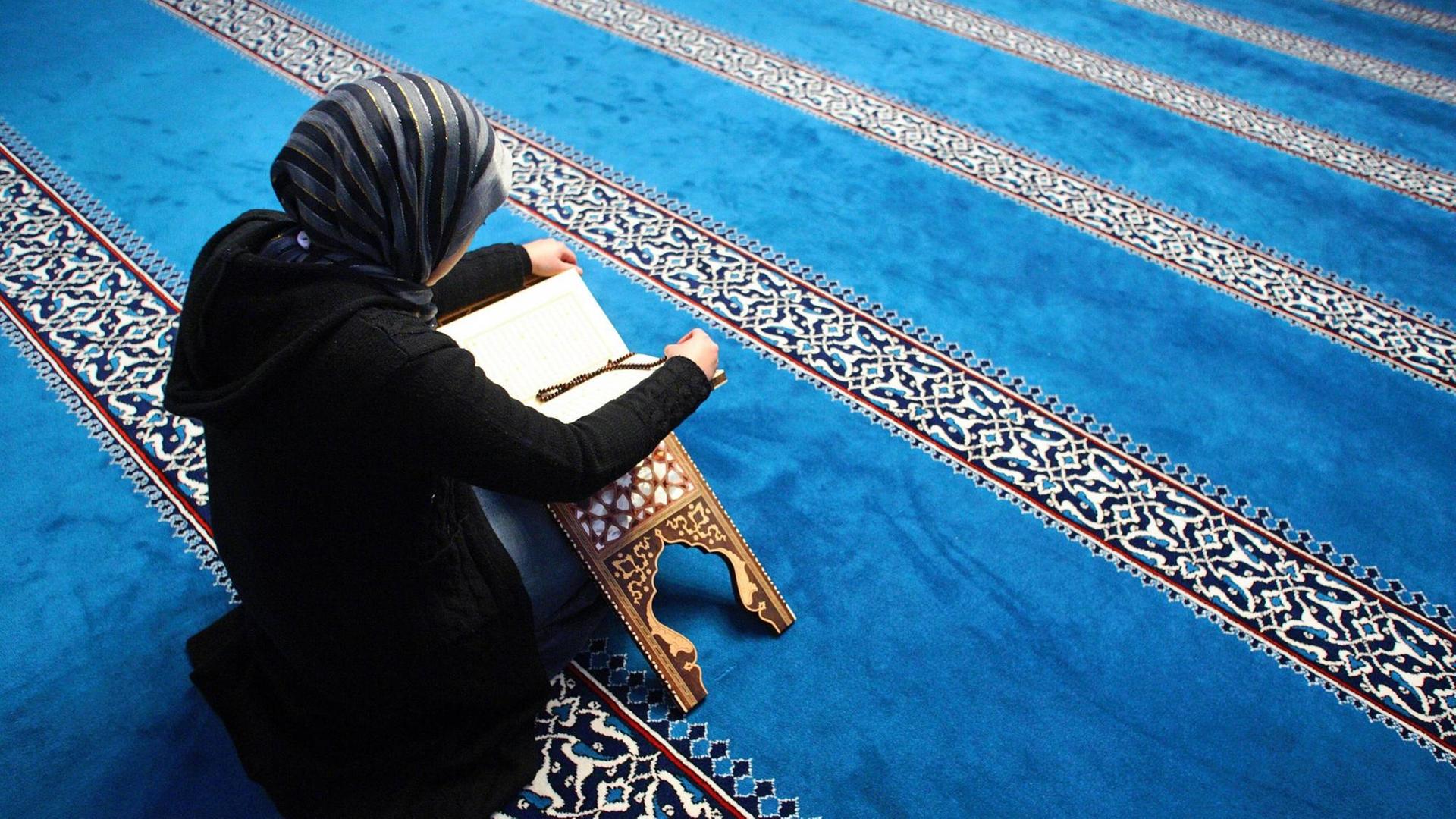 Eine Frau liesst in der Sehitlik Moschee am Columbiadamm in Berlin-Neukölln im Koran. Sie ist schwarz gekleidet, der Teppich in der Moschee ist überwiegend blau. Eine Frau liesst in der Sehitlik Moschee am Columbiadamm in Berlin-Neukölln im Koran. Sie ist schwarz gekleidet, der Teppich in der Moschee ist überwiegend blau.