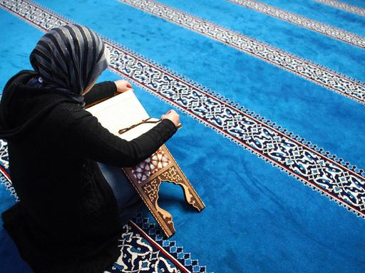 Eine Frau liesst in der Sehitlik Moschee am Columbiadamm in Berlin-Neukölln im Koran. Sie ist schwarz gekleidet, der Teppich in der Moschee ist überwiegend blau.