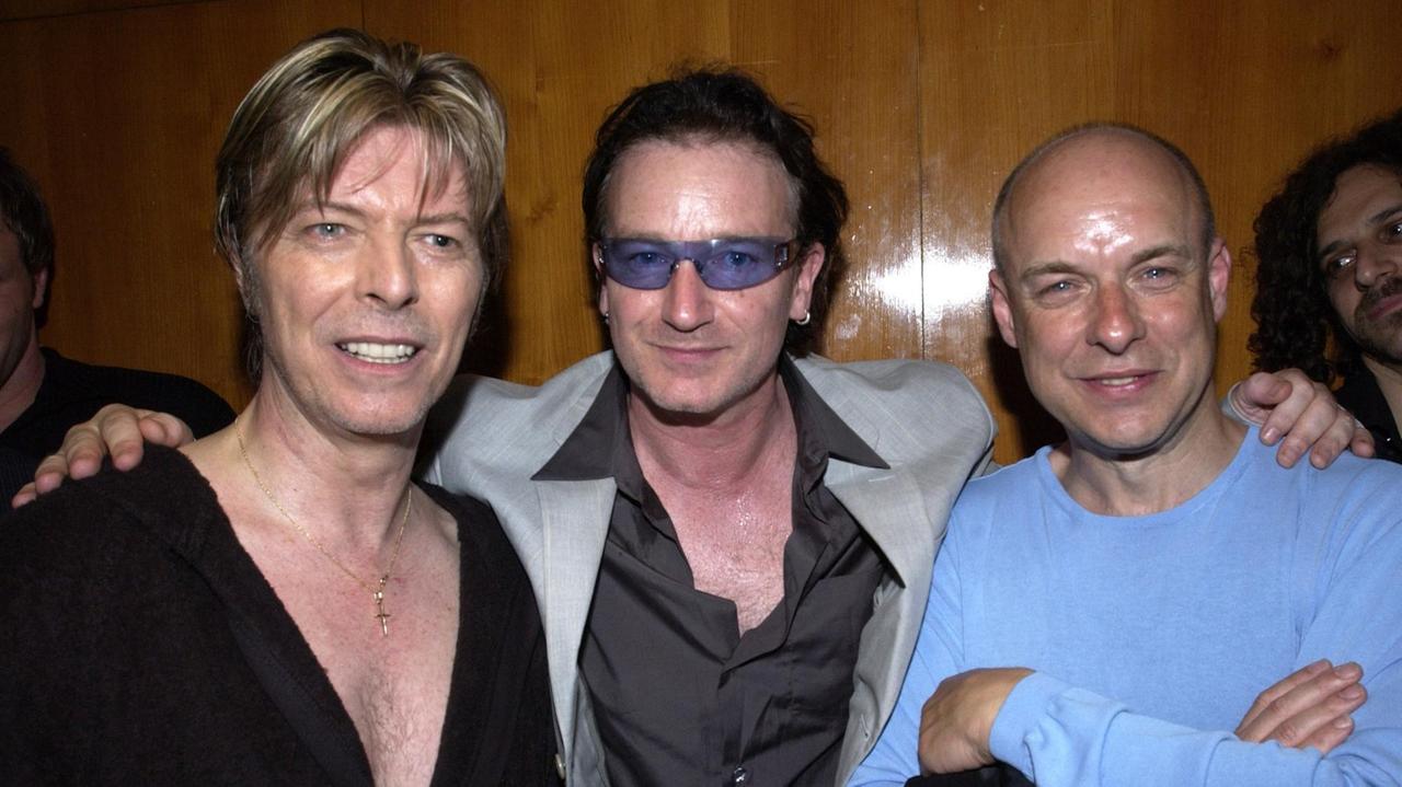 David Bowie, Bono und Brian Eno gemeinsam auf einem Foto von 2002. David Bowie, Bono und Brian Eno gemeinsam auf einem Foto von 2002.