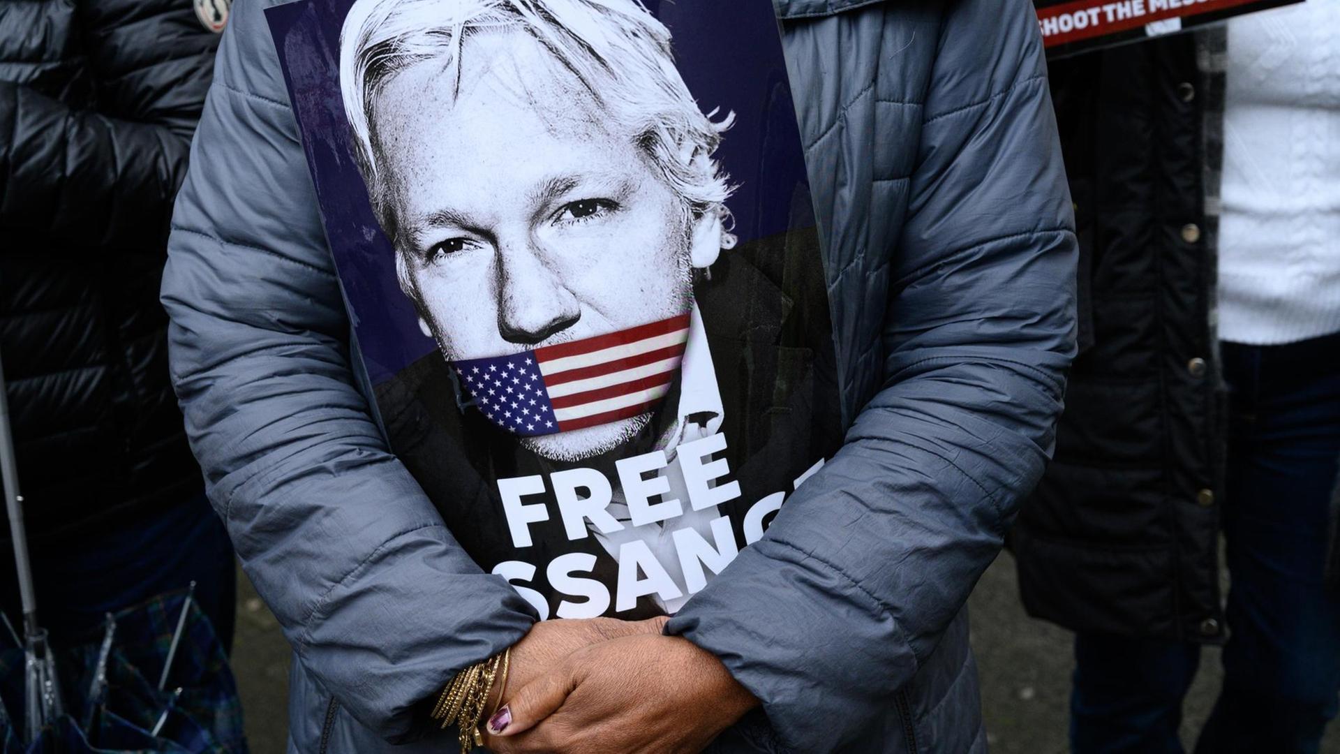 Eine Unterstützerin von Julian Assange hält vor ihrer Brust ein Schild, auf dem das Gesicht von Assange zu sehen ist. Der Mund von Assange ist auf dem Bild mit einer US-amerikanischen Flagge zugeklebt. Darunter steht auf dem Plakat "Free Assange" ("Befreit Assange").