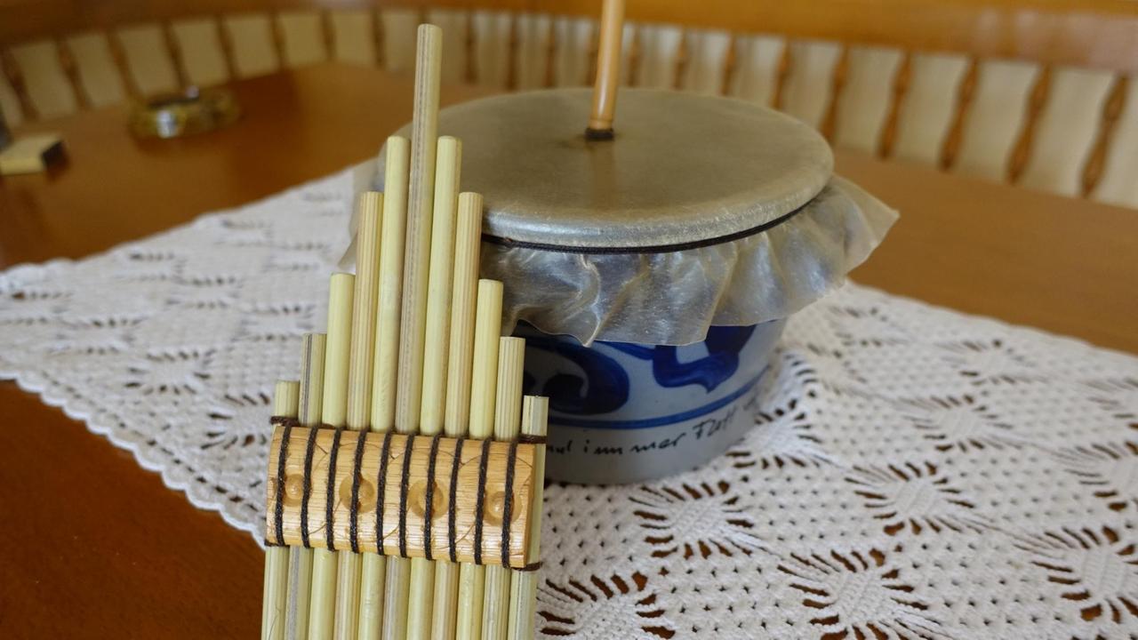 Eine Schüssel mit Lederdeckel und eine Panflöte sind auf einem Küchentisch mit Häkeldecke drappiert. Eine Schüssel mit Lederdeckel und eine Panflöte sind auf einem Küchentisch mit Häkeldecke drappiert.