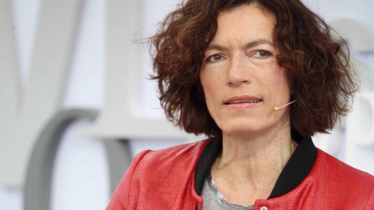 Anne Weber hat dunkle, lockige Haare und trägt eine rote Jacke und ein graues Shirt. Im Hintergrund sind große, ineinander verschachtelte Buchstaben an einer Wand zu sehen. Anne Weber hat dunkle, lockige Haare und trägt eine rote Jacke und ein graues Shirt. Im Hintergrund sind große, ineinander verschachtelte Buchstaben an einer Wand zu sehen.
