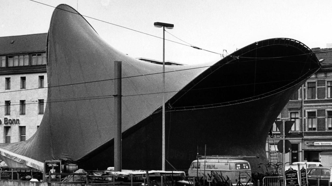 Schwarz-Weiß-Foto eines ausgeprochen geschwungenen Zeltes, an dem viele Autos vorbei fahren. Schwarz-Weiß-Foto eines ausgeprochen geschwungenen Zeltes, an dem viele Autos vorbei fahren.