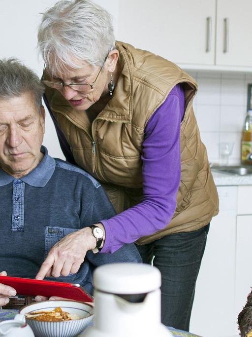 Vier ältere Menschen beim gemeinsamen Kaffeetrinken in der Küche schauen auf ein rotes Tablet