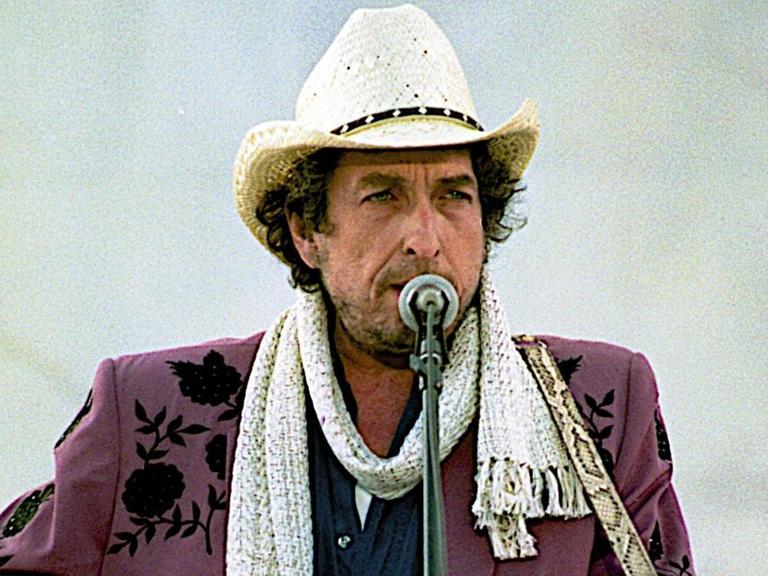 Bob Dylan vor einem Mikrofon. Er rtägt eine rotbraune Jacke, einen weißen Schal und einen weißen Hut.