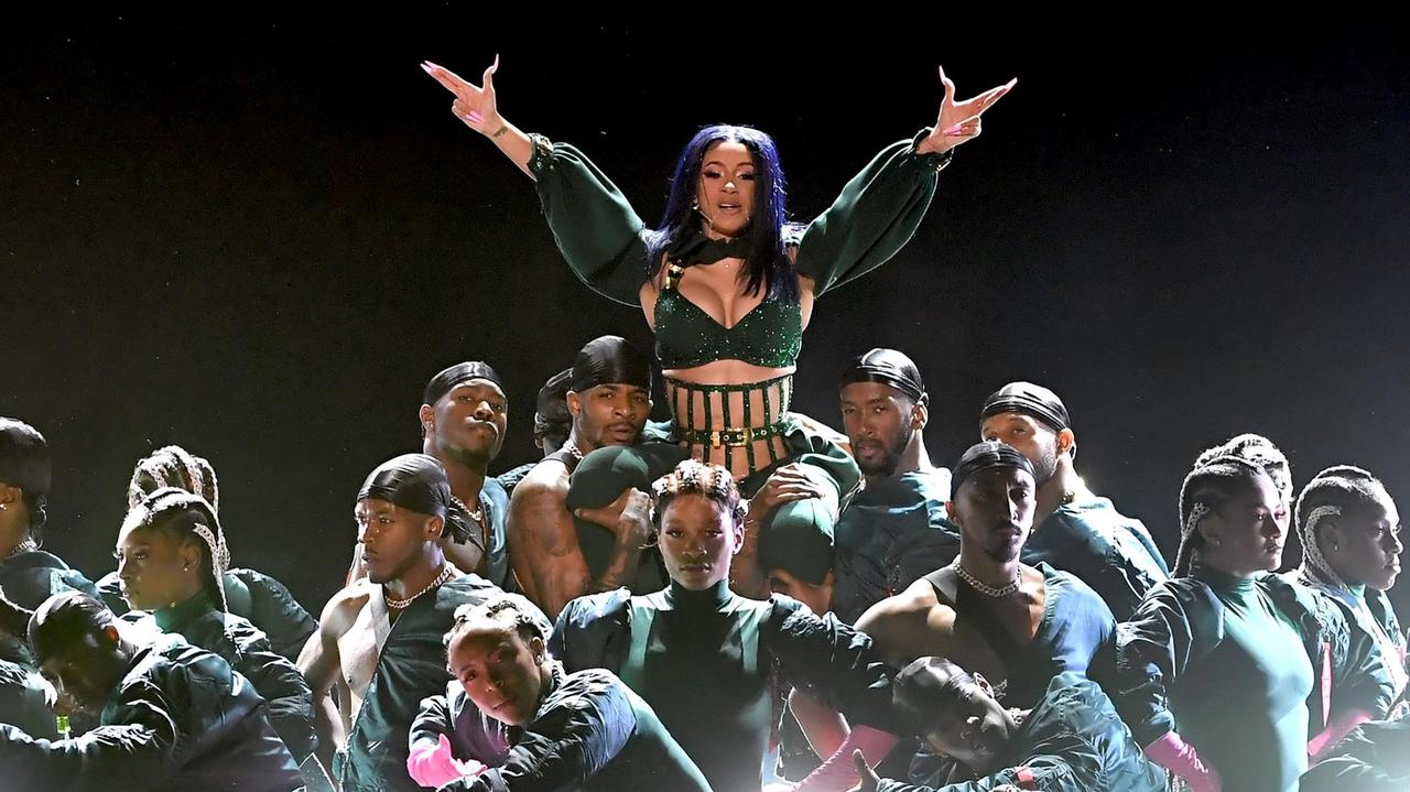 Das Bild zeigt Cardi B während eines Auftritts. Sie wird von einer Gruppe von Männern getragen. Ihre beiden Arme hat sie in die Luft gestreckt und ihre Hände zu imaginären Waffen geformt. Das Bild zeigt Cardi B während eines Auftritts. Sie wird von einer Gruppe von Männern getragen. Ihre beiden Arme hat sie in die Luft gestreckt und ihre Hände zu imaginären Waffen geformt.