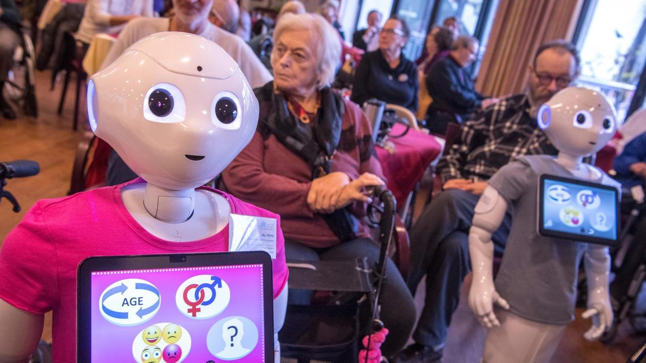 Zwei menschenähnliche Roboter stehen mit bunten T-Shirts zwischen älteren Menschen im Altenpflegeheim Zwei menschenähnliche Roboter stehen mit bunten T-Shirts zwischen älteren Menschen im Altenpflegeheim