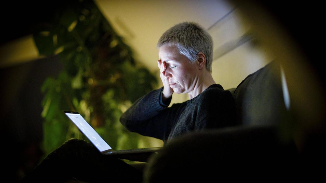 Eine Frau sitzt mit geschlossenen Augen auf dem Sofa vor ihrem Laptop und hat ihre Hand auf die Stirn gelegt. Symbolbild.