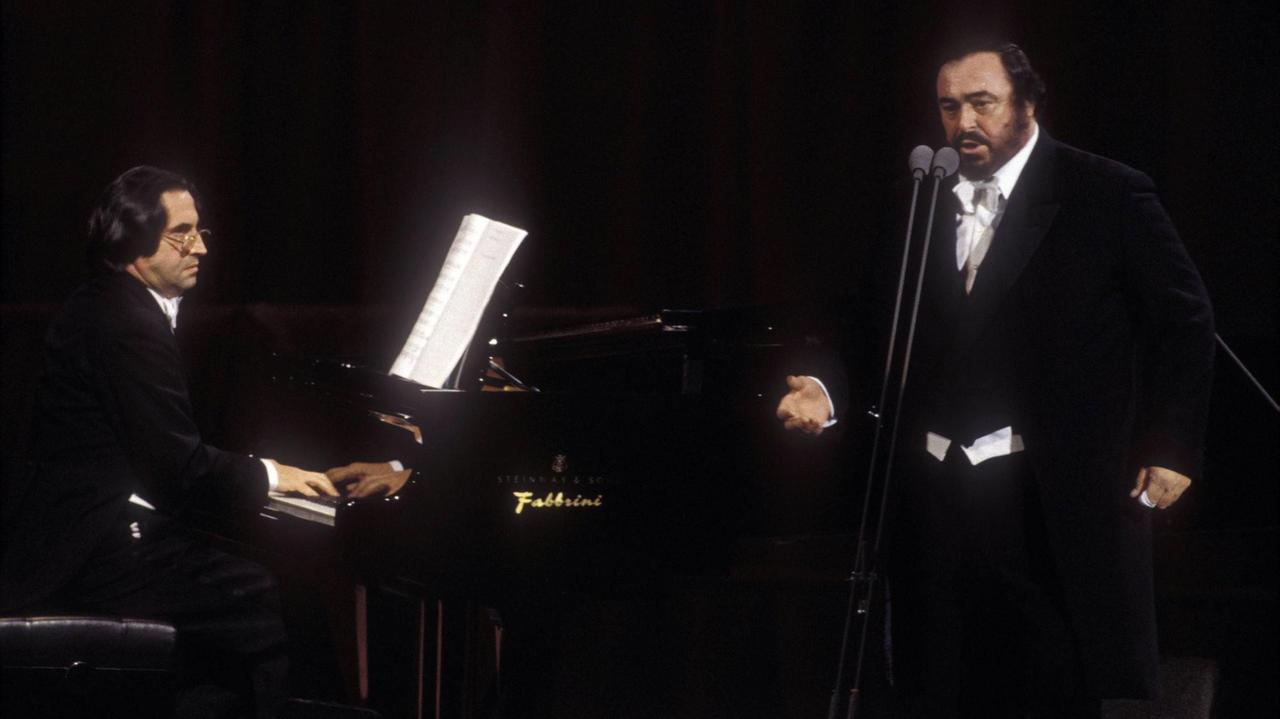 Riccardo Muti sitzt mit Brille am Klavier, den Startenor Luciano Pavarotti begleitend, der hinter einem Mikrophonpaar steht.