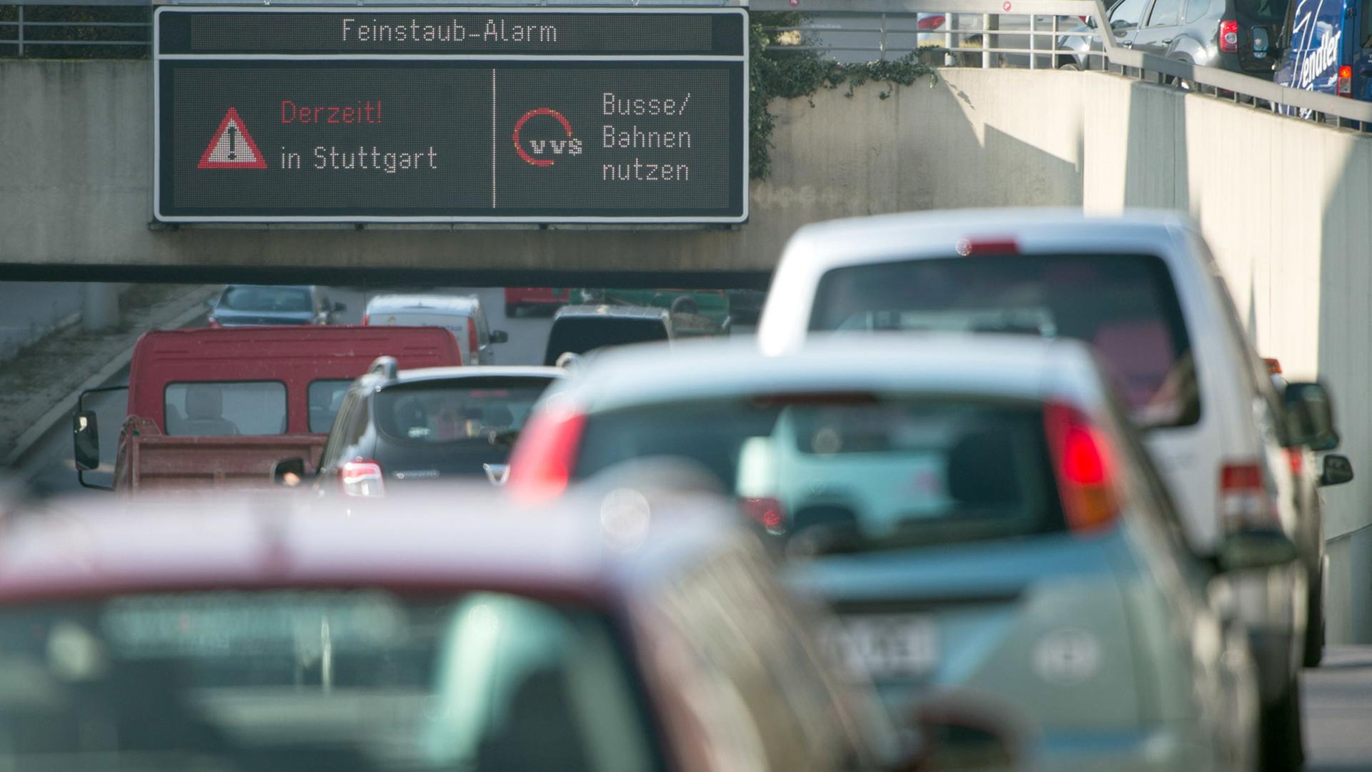 Autos fahren in Stuttgart durch die Innenstadt, während auf einer Anzeige ein Feinstaub-Alarm für die Umweltzone Stuttgart angezeigt und auf öffentliche Verkehrsmittel hingewiesen wird.