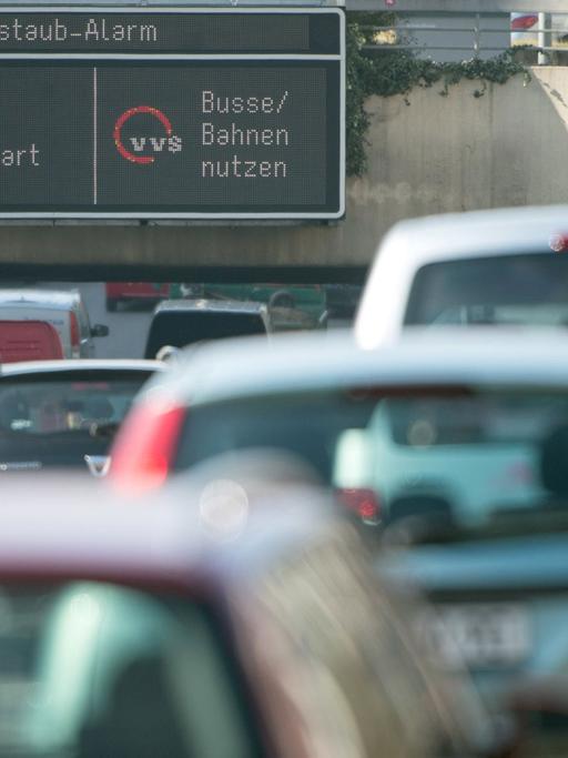 Autos fahren in Stuttgart durch die Innenstadt, während auf einer Anzeige ein Feinstaub-Alarm für die Umweltzone Stuttgart angezeigt und auf öffentliche Verkehrsmittel hingewiesen wird.