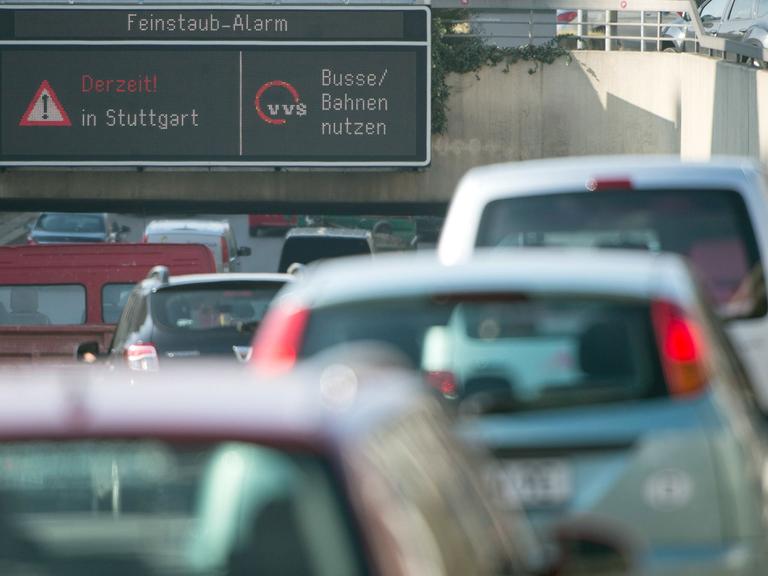 Autos fahren in Stuttgart durch die Innenstadt, während auf einer Anzeige ein Feinstaub-Alarm für die Umweltzone Stuttgart angezeigt und auf öffentliche Verkehrsmittel hingewiesen wird.