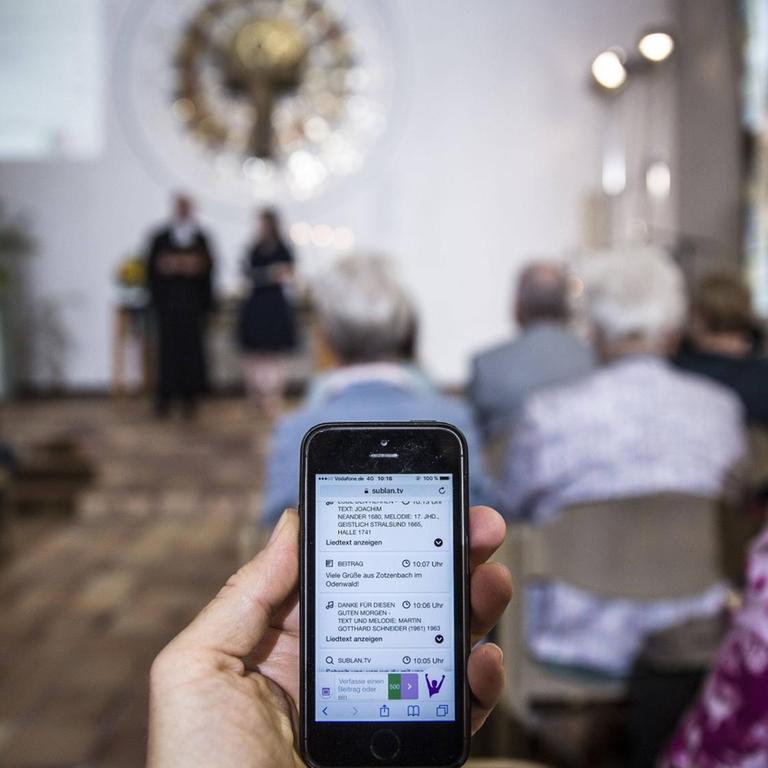 Bei einem Gottesdienst im Rahmen des "Sublan"-Projektes im Juli 2017 in Hessen können sich die Besucher per Smartphone und Tablet-Computer direkt in die Predigt und die Fürbitte-Gebete einschalten. Im Vordergrund hält eine Person ein Smartphone, im Hintergrund ist der Gottesdienst zu sehen.