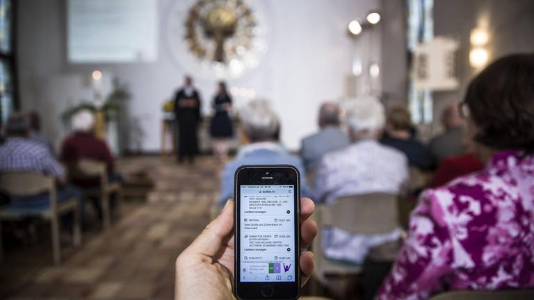 Bei einem Gottesdienst im Rahmen des "Sublan"-Projektes im Juli 2017 in Hessen können sich die Besucher per Smartphone und Tablet-Computer direkt in die Predigt und die Fürbitte-Gebete einschalten. Im Vordergrund hält eine Person ein Smartphone, im Hintergrund ist der Gottesdienst zu sehen.